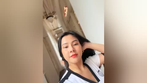 Media: Video of an Asian woman with straight black hair, fair skin, and red lipstick, wearing a white shirt with black accents, standing in a dimly lit hallway.