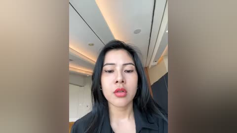 Media: Video of an Asian woman with long black hair, wearing a black jacket, standing indoors with a modern, minimalist interior. The background features white walls, recessed lighting, and a neutral color palette.