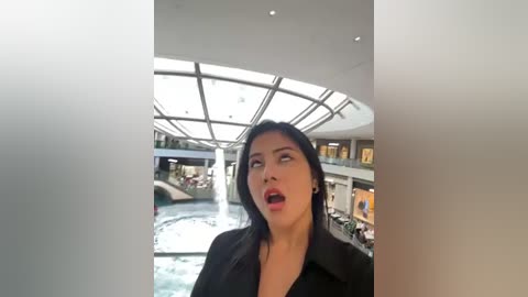 Media: Video of a woman with medium skin tone, long black hair, wearing a black jacket, in a modern indoor space with a glass ceiling and a pool in the background.