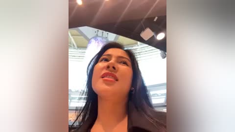 Media: Video of a smiling woman with long black hair, wearing a black top, taken from a low angle inside a modern, well-lit building with large windows and a chandelier.