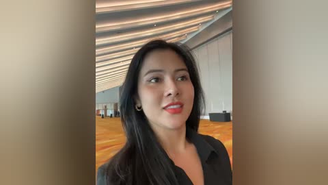 Media: Video of a young Asian woman with long black hair and fair skin, wearing a black blazer, standing in a modern, brightly lit room with a wooden floor and sleek, white walls.