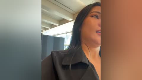 Media: Video of a woman with medium brown skin, wearing a black blazer, partially obscured by a beige wall, in an office with modern architecture.