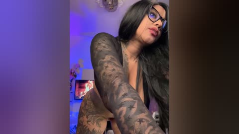 Media: Video of a tattooed woman with long black hair, wearing black-framed glasses, standing in a dimly lit room with blue lighting. She's topless, showcasing extensive arm and chest tattoos.