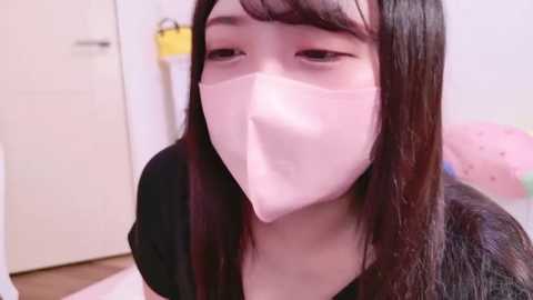 Media: Video of a young East Asian woman with long, straight black hair and fair skin, wearing a pink surgical mask, a black top, and standing indoors with a white door and a yellow object in the background.