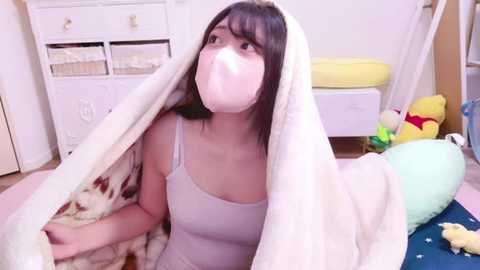 Media: Video of a young woman with shoulder-length black hair, wearing a white mask, lying under a fluffy white blanket on a pink carpet in a bright, tidy room with toys and a white dresser in the background.