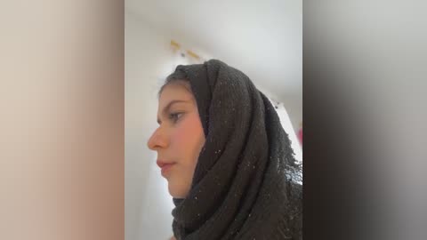 Media: Video of a woman with fair skin, wearing a black lace hijab, facing left in a side profile. Background is a blurred, light-colored wall.