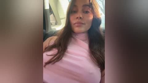 Media: A video of a young woman with long, straight, dark brown hair, wearing a pink, sleeveless turtleneck sweater. She is seated in a car with the sun streaming in. Her expression is neutral, and the background is blurred.