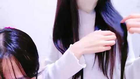 Media: Video of a woman with long, straight black hair, wearing a white sweater, holding a hairbrush, brushing her hair. Background is blurred, focusing on the hair and brushing action.