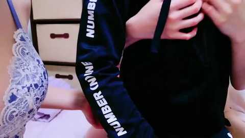 Media: Video of a woman wearing a black sweater with white \"NUMBER ONE\" text, holding a black handbag, against a backdrop of beige furniture and a lace bra.