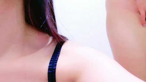 Media: A close-up video of a woman with fair skin and dark hair, wearing a black lace bra, showing her bare shoulder and upper chest. The background is a plain, light-colored wall, emphasizing the texture of the lace and her smooth skin.