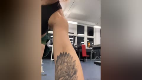 Media: A video captures a woman's side profile in a gym, wearing a black sports bra and revealing a detailed feather tattoo on her lower left side. The background features gym equipment and large windows.