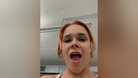 Media: Video of a woman with fair skin and red hair, wearing a beige top, mouth open in an exaggerated expression, in a white, slightly messy room with a ceiling light.