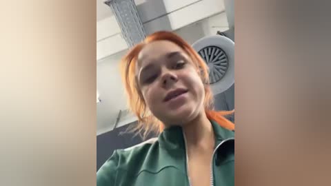 Media: Video of a young woman with light skin and red hair, wearing a green jacket, in an office setting with a ceiling fan and gray walls.