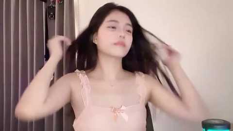 Media: Video of an Asian woman with long black hair, light skin, wearing a pink lace camisole, adjusting her hair, against a beige wall with vertical blinds.