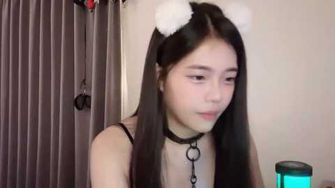 Media: Video of an Asian woman with long black hair wearing white bear ears and a black choker, sitting in a room with vertical blinds and a green lamp.