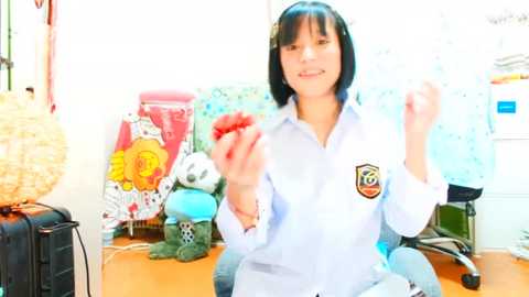 Media: Video of an Asian woman with black hair, wearing a light blue shirt, playing with a toy in a cluttered office with colorful stuffed animals, papers, and a desk lamp.