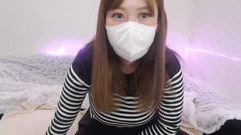 Media: Video of an Asian woman with long brown hair, wearing a white mask, black-and-white striped shirt, and black pants, kneeling on a bed with a white fluffy blanket, soft purple LED lights in the background.