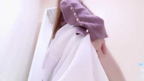 Media: A video captures a person wearing a mauve ribbed sweater, lifting a white, textured garment, revealing a smooth white background. The image is soft-focused, emphasizing the texture contrast between the sweaters.