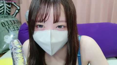 Media: Video of an East Asian woman with long, straight brown hair, wearing a white face mask, blue tank top, and purple pillow. Background features a pink curtain and a white \"Cleaner\" bag.