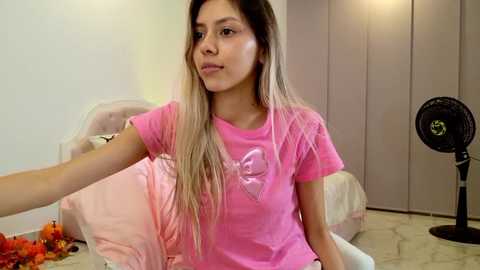 Media: Video of a young Asian woman with long blonde hair in a pink T-shirt, sitting on a bed with a fan and colorful flowers on the floor.