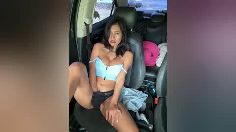 Media: Video of a young Asian woman with long black hair, wearing a blue bra and black shorts, sitting provocatively in a car, legs spread, surrounded by clothes and a pink pillow.