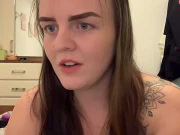 Media: Video of a young woman with fair skin, long brown hair, and a floral tattoo on her shoulder, wearing a pink top, in a bathroom with white cabinets and a pink towel hanging.