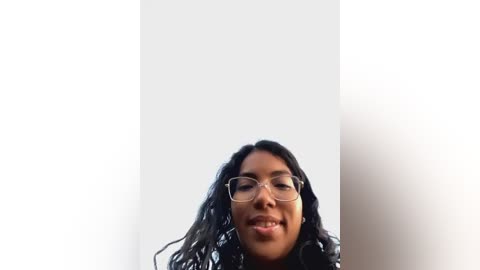 Media: Video of a smiling, dark-skinned woman with curly hair and glasses, standing against a stark white background. Her expression is cheerful, and she appears to be wearing a black top.