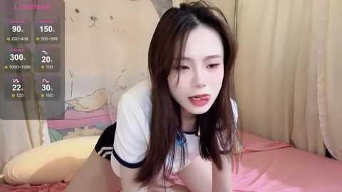 Media: Video of an East Asian woman with long, straight brown hair, fair skin, and light makeup, wearing a white shirt and black shorts, lying on a pink bed with a cartoon print.