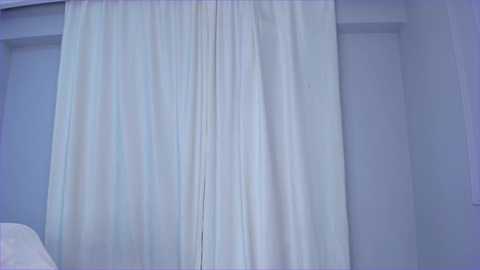 Media: Video of a minimalist bedroom featuring a large, central, sheer white curtain draped over a window. The background wall is light blue, creating a serene and calming atmosphere.