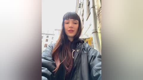 Media: Video of a young woman with long, straight, dark brown hair and blunt bangs, wearing a black leather jacket, standing in an urban street with light-colored buildings in the background.