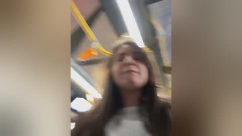 Media: Video of a young woman with long, dark hair and light skin, wearing a grey top and jacket, smiling in a brightly lit subway car with yellow poles and overhead lights.