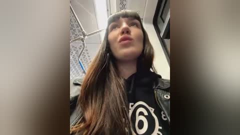 Media: Video of a young woman with long brown hair, wearing a black hoodie with a white number 6 design, looking up from a subway seat.