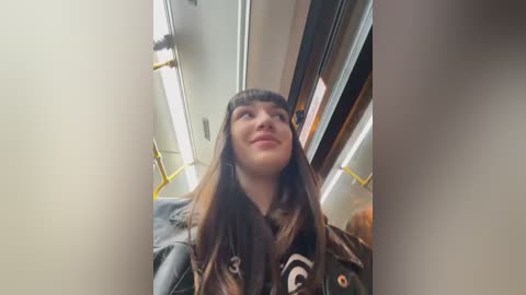 Media: A video of a young woman with long brown hair, wearing a black jacket, smiling inside a bus. The background shows yellow handrails and fluorescent lights.