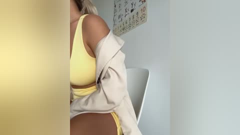 Media: A video of a woman in a yellow bra and matching panties, wearing a beige cardigan, sitting on a white chair in a minimalist room with a calendar on the wall.