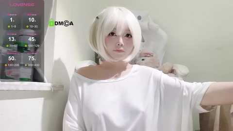 Media: Video of a young woman with pale skin, short platinum blonde hair, wearing a loose white t-shirt, standing in a well-lit room with a window, holding a phone.