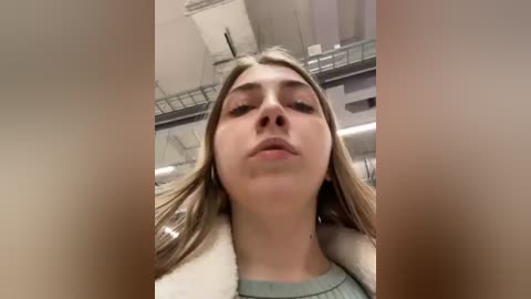 Media: Video of a young woman with fair skin, straight blonde hair, wearing a light green sweater, standing indoors with a blurry, industrial background.