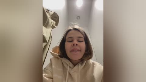 Media: Video of a young woman with straight, shoulder-length brown hair, wearing a beige hoodie, looking up with a playful expression. She is inside a narrow, bright space with a ceiling light.