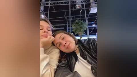 Media: Video of two young women, one with light skin and straight hair, the other with darker skin and curly hair, lying close together on a black leather couch. Background features a modern, industrial-style room with metallic structures and green plants.