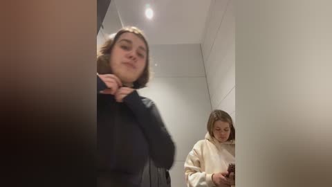 Media: Video of a young woman with shoulder-length hair in a black coat adjusting her collar, standing in a dimly lit bathroom with white tiled walls. Another young woman in a beige sweater stands in the background, texting on her phone.