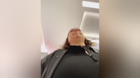 Media: Video of a woman with medium-length brown hair, wearing a black jacket, standing in an elevator. The background features a minimalist, modern interior with white walls and fluorescent lighting. The image is slightly blurred, giving it a dreamy, surreal feel.