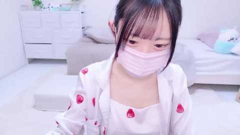 Media: Video of an Asian woman in a white shirt with strawberry patterns, wearing a pink face mask, sitting in a light-colored bedroom with a white dresser and bed.