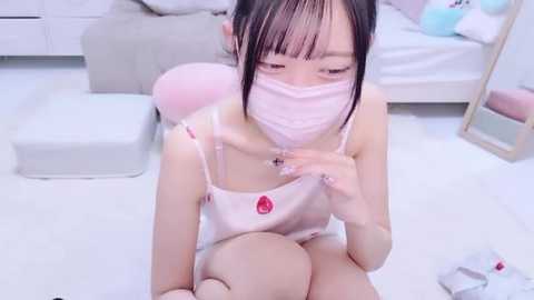 Media: A video of an East Asian woman with fair skin and black hair, wearing a pink face mask, camisole, and underwear, sitting on a white floor with a stuffed animal.