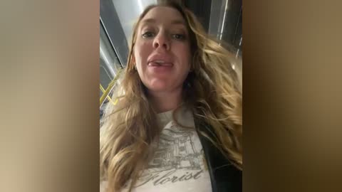 Media: Video of a blonde woman with long hair, wearing a white \"Harvest\" t-shirt, leaning forward in a lift, tongue sticking out.