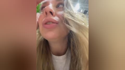 Media: Video of a young blonde woman with straight hair, wearing a white shirt, tilted head, and a playful expression, captured from a low angle.