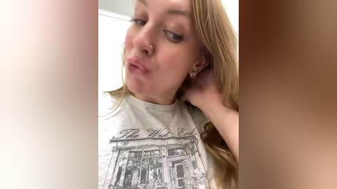 Media: A video of a young woman with light skin, long blonde hair, and a septum piercing, making a kissy face while wearing a graphic T-shirt depicting a storefront.