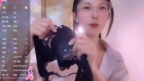 Media: Video of an East Asian woman with fair skin, straight black hair, and glasses, holding a black lace bra. She's wearing a light blouse and has a neutral expression.