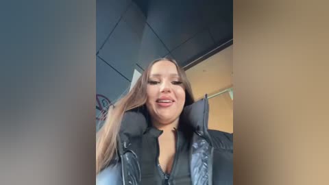 Media: Video of a young Caucasian woman with long brown hair, wearing a black jacket, seated in a car with a dark interior and muted background.