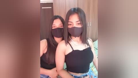 Media: Video of two Asian women with medium skin tones, black masks, and black tank tops, sitting side by side. One wears a floral skirt, the other a denim skirt. Background shows wooden wardrobes and a bed.