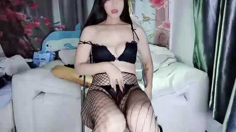 Media: Video of a pale-skinned, long-haired Asian woman with black lingerie, fishnet stockings, and a black bra, sitting on a white couch in a room with pink floral wallpaper and a blue horse toy.