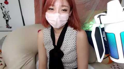 Media: Video of an Asian woman with red hair, wearing a black tie, face mask, and patterned top, seated on a white couch, surrounded by colorful artwork and a blue-and-white speaker.